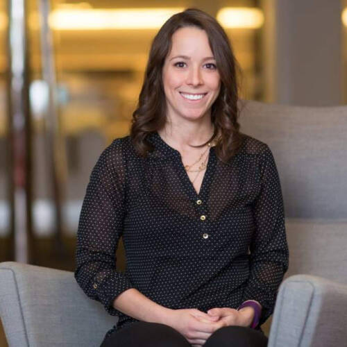 A woman sitting on a gray chair, smiling, wearing a black blouse with small white dots, in a warmly lit indoor setting.
